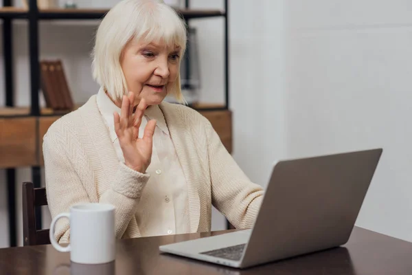 Senior woman sitting at computer desk and waving while having video call at home — Stock Photo