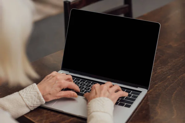 Cropped view of senior woman sitting at table and typing on laptop with blank screen at home — Stock Photo