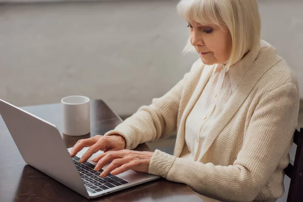 Старшая женщина сидит за столом и печатает на ноутбуке дома — стоковое фото
