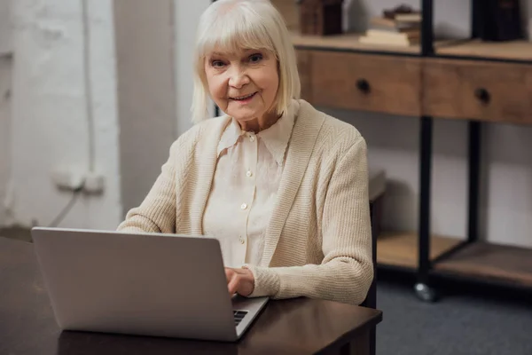Mujer mayor sonriente sentada en la mesa y escribiendo en el ordenador portátil en casa - foto de stock