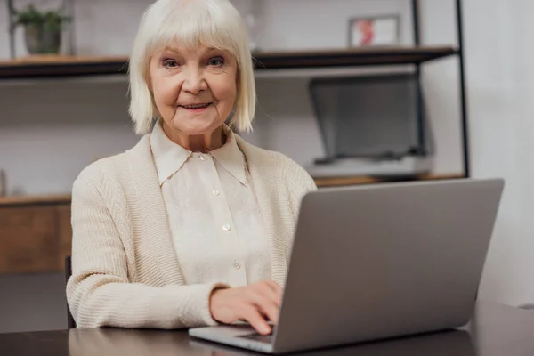 Smiling senior woman sitting at table, looking at camera and typing on laptop at home — Stock Photo