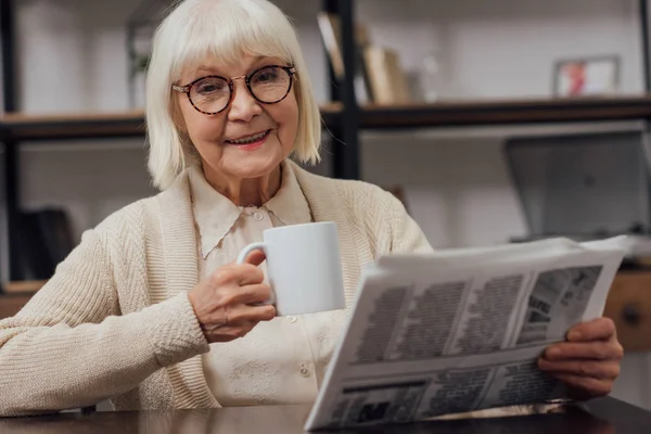 Щаслива старша жінка сидить за столом і читає газету під час пиття кави вдома — стокове фото