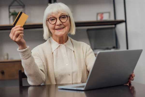 Mulher sênior em óculos sentado na mesa do computador, segurando cartão de crédito e fazendo compras on-line em casa — Fotografia de Stock