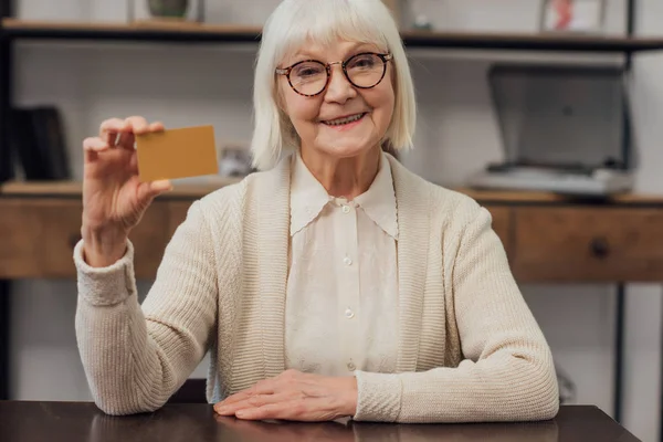 Felice donna anziana seduta a tavola, guardando la fotocamera e tenendo la carta di credito a casa — Foto stock