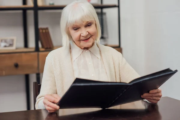 Sonriente mujer mayor con el pelo gris sentado en la mesa y mirando álbum de fotos en casa - foto de stock