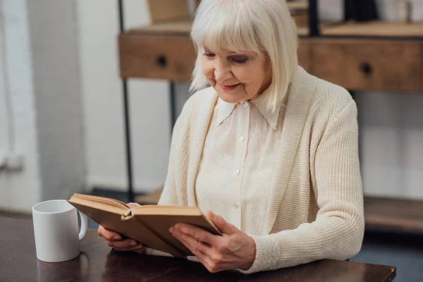 Lächelnde Seniorin sitzt zu Hause am Tisch und liest Buch — Stockfoto