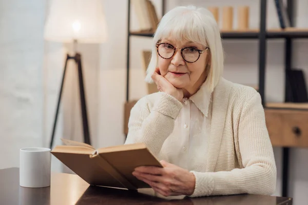 Donna anziana in bicchieri seduta a tavola, mento appoggiato con mano e libro di lettura a casa — Foto stock