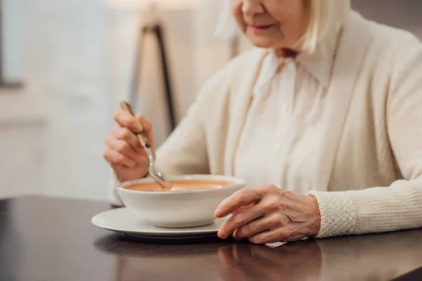 Cropped view of senior woman sitting and eating cream soup at home — Stock Photo
