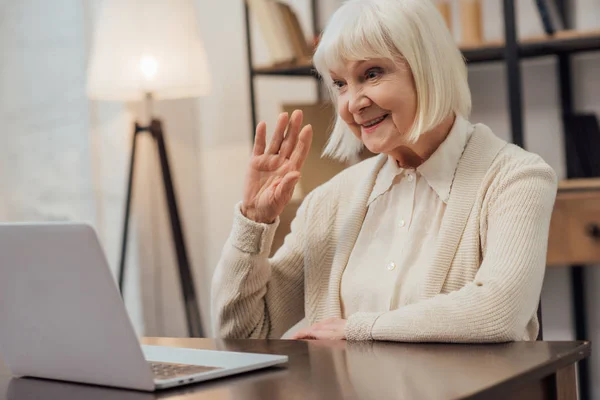 Улыбающаяся пожилая женщина сидит за компьютерным столом и машет рукой во время видеозвонка дома — стоковое фото