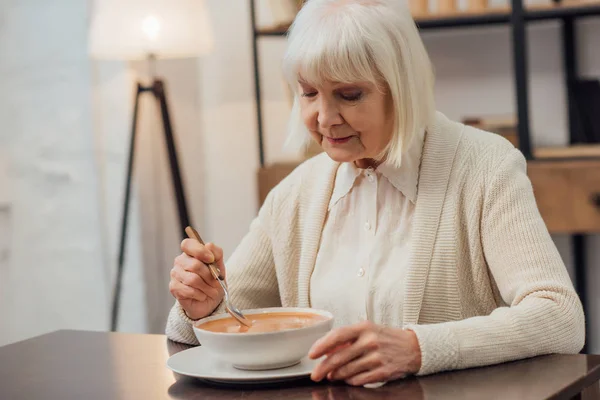 Foco seletivo da mulher idosa sentada à mesa e comendo sopa de creme em casa — Fotografia de Stock