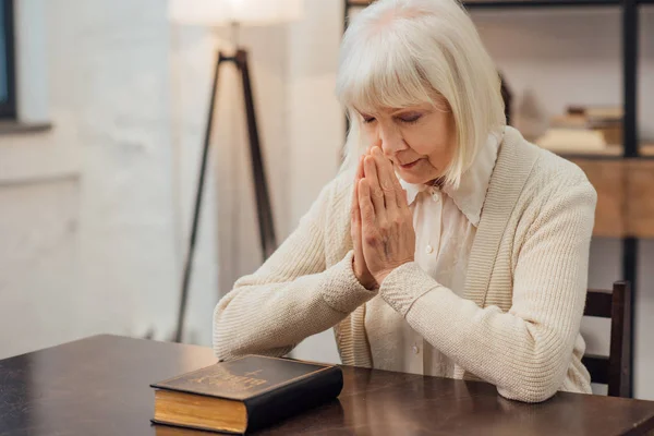 Konzentrierte Seniorin sitzt zu Hause vor heiliger Bibel und betet — Stockfoto
