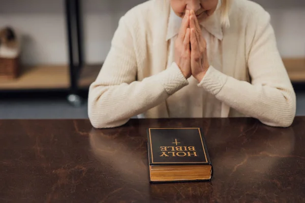 Cropped view of senior woman sitting and praying in front of holy bible at home — Stock Photo