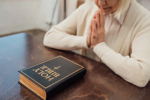 Partial view of senior woman sitting and praying in front of holy bible at home — Stock Photo