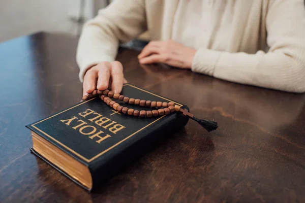Обрезанный вид пожилой женщины, сидящей за столом с четками и святой библией дома — стоковое фото