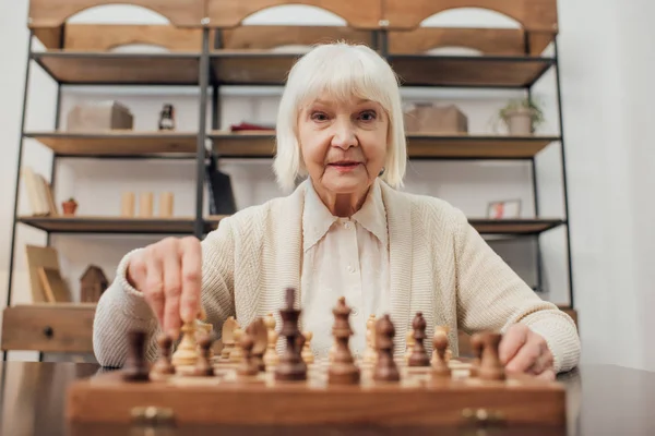 Senior woman sitting at table, looking at camera and playing chess at home — Stock Photo