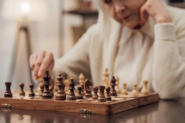 Vista cortada da mulher idosa sentada à mesa, apoiando o queixo com a mão e jogando xadrez em casa — Fotografia de Stock