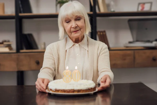 Traurige Seniorin mit grauen Haaren sitzt zu Hause mit Geburtstagstorte am Tisch — Stockfoto