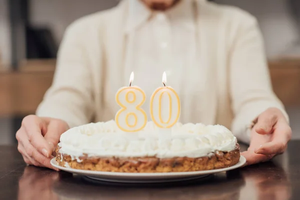 Visão parcial da mulher idosa segurando bolo de aniversário com o número 80 no topo em casa — Fotografia de Stock