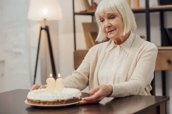 Mujer mayor con el pelo gris sentado y la celebración de la torta con el número 80 en la parte superior mientras se celebra el cumpleaños en casa — Stock Photo