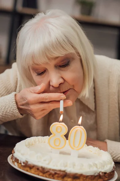 Senior woman lighting up cigarette from burning candles on birthday cake at home — Stock Photo