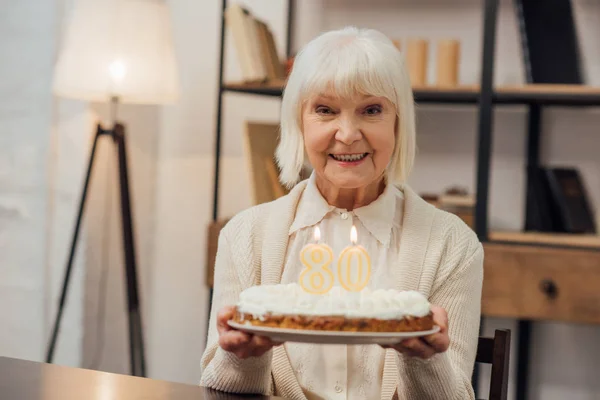 Donna anziana sorridente che tiene la torta con il numero 80 in cima e festeggia il compleanno a casa — Foto stock