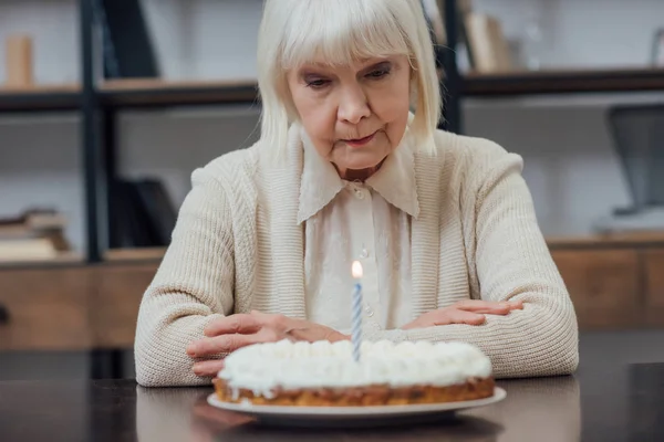 Mujer mayor disgustada sentada en la mesa y mirando pastel de cumpleaños con vela encendida en casa - foto de stock