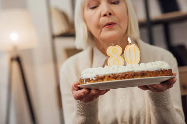 Mujer mayor soplando velas en la torta con el número 80 en la parte superior mientras se celebra el cumpleaños en casa — Stock Photo