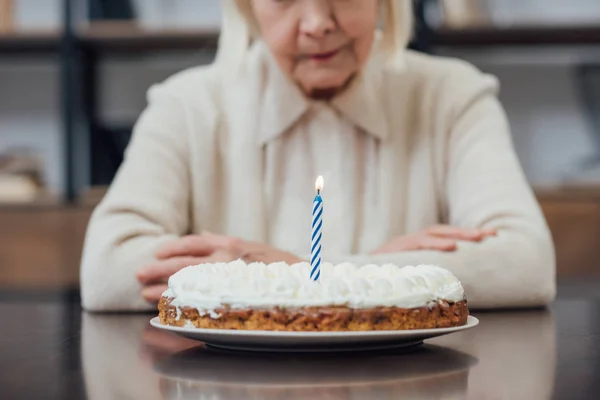 Vista cortada da mulher sênior sentada à mesa e olhando para o bolo de aniversário com vela acesa em casa — Fotografia de Stock