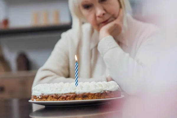 Selektiver Fokus trauriger Seniorin, die am Tisch sitzt und zu Hause Geburtstagstorte mit brennender Kerze betrachtet — Stockfoto
