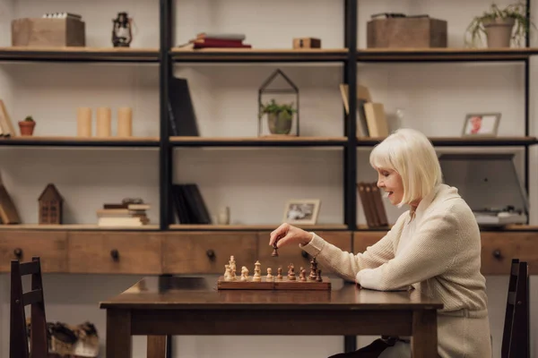 Mulher sênior sentada à mesa e jogando xadrez na sala de estar — Fotografia de Stock