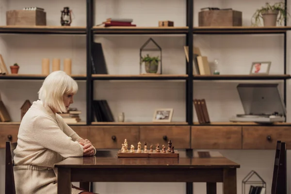 Грустная старшая женщина сидит за столом с шахматной игрой в гостиной — стоковое фото