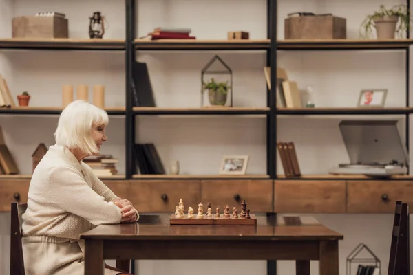 Senior femme aux cheveux gris assis à table avec jeu d'échecs dans le salon — Photo de stock