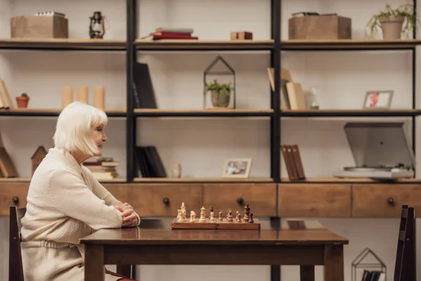 Anciana sola sentada en la mesa con juego de ajedrez en la sala de estar - foto de stock