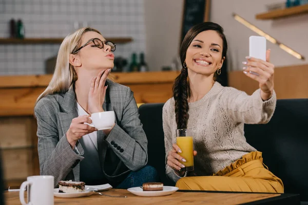 Cheerful women taking selfie and holding drinks in cafe — Stock Photo