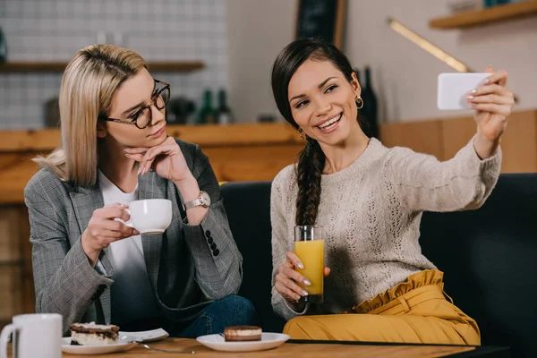 Mujeres hermosas tomando selfie y sosteniendo bebidas en la cafetería - foto de stock