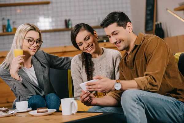 Bel homme tenant smartphone près des femmes attrayantes dans le café — Photo de stock