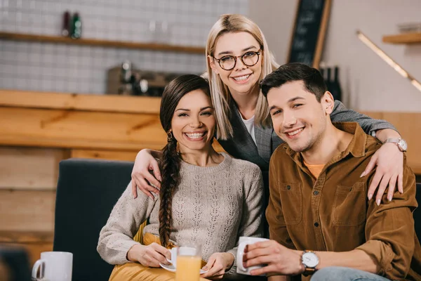 Веселая женщина в очках обнимает улыбающихся друзей в кафе — стоковое фото