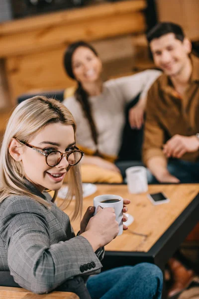 Selective focus of attractive woman in glasses holding cup with friends on background — Stock Photo