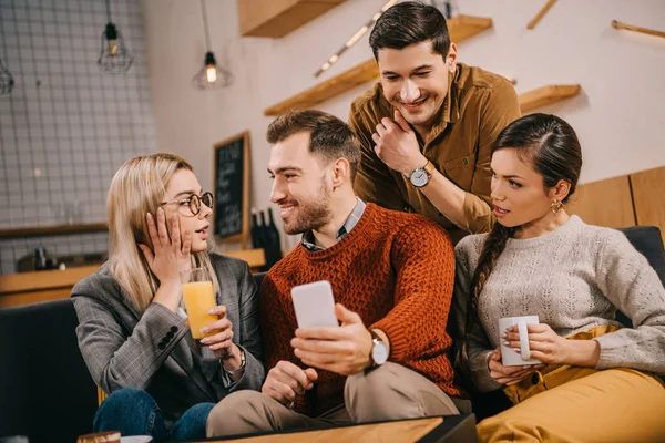 Mujer sorprendida mirando al hombre sosteniendo teléfono inteligente cerca de amigos - foto de stock