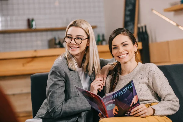 Atractiva mujer sosteniendo la revista de belleza y sonriendo amigo cercano en gafas - foto de stock