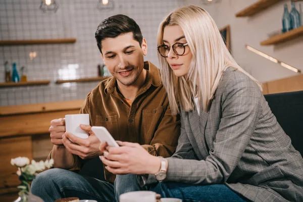 Женщина в очках держит смартфон рядом с мужчиной с чашкой — стоковое фото