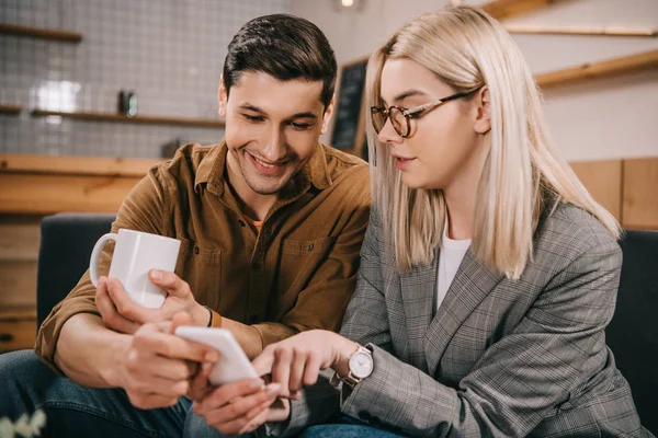 Mujer en gafas apuntando con el dedo al teléfono inteligente cerca de hombre con taza - foto de stock