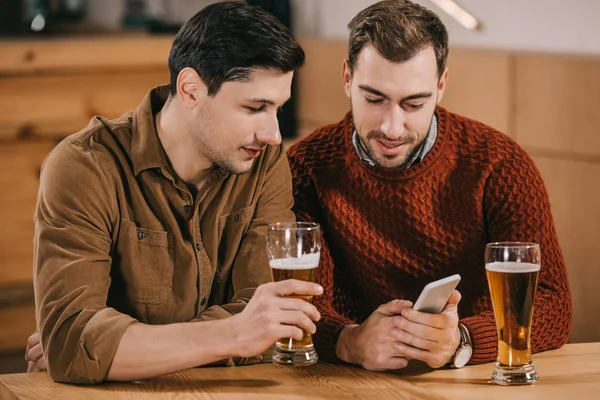 Hombres guapos mirando el teléfono inteligente cerca de vasos con cerveza - foto de stock