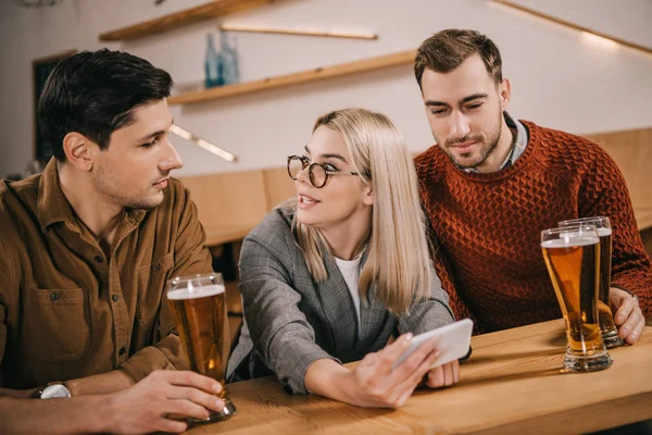 Sorprendida mujer sosteniendo teléfono inteligente cerca de amigos varones con vasos de cerveza - foto de stock