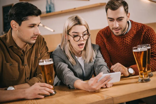 Здивована жінка дивиться на смартфон поруч з друзями-чоловіками в келихах пива — стокове фото