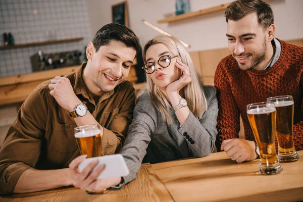 Atractiva mujer tomando selfie con amigos masculinos cerca de vasos de cerveza - foto de stock