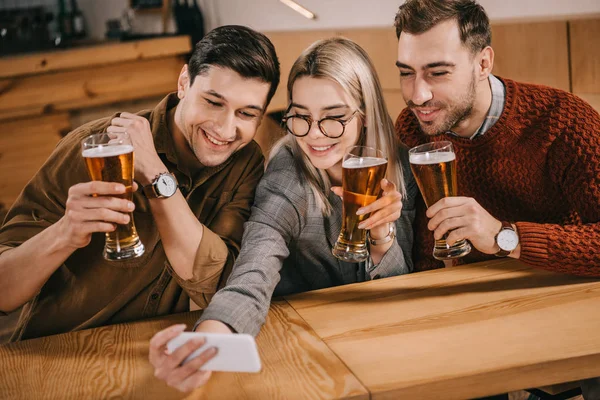 Hermosa mujer tomando selfie con amigos masculinos y sosteniendo vaso de cerveza - foto de stock