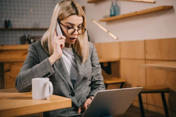 Messa a fuoco selettiva della donna scioccata in occhiali che parla sullo smartphone mentre guarda il computer portatile — Foto stock