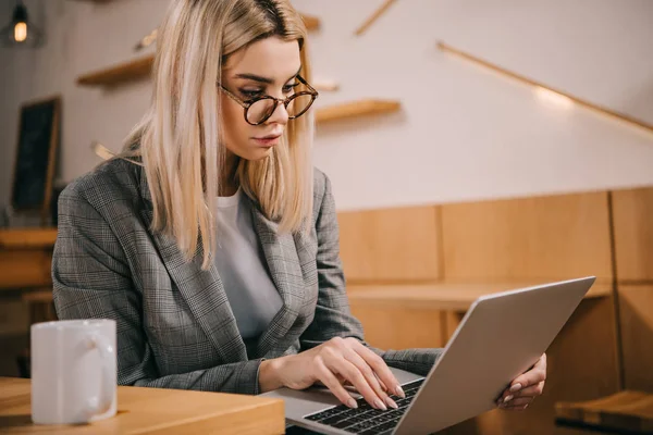 Foco seletivo da mulher atraente em óculos digitando no laptop no café — Fotografia de Stock