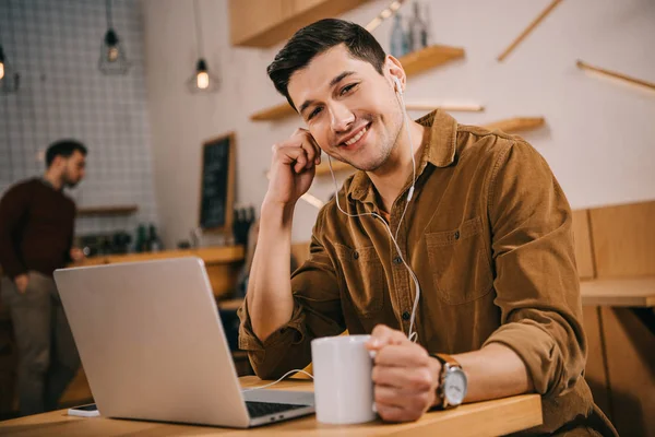 Schöner Mann mit Kopfhörer, der eine Tasse Kaffee in der Hand hält und im Café in die Kamera schaut — Stockfoto